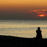 A silhouetted figure sits alone on beach at sunset